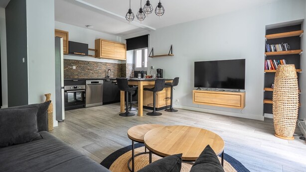 A modern kitchen with a stone backsplash, a dining area with a wooden table, and a living room with a sofa and a TV. - L’Escapade du Quai - Proche Gare (Pontivy)
