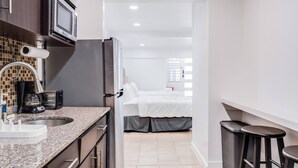 Una cocina moderna con encimeras de granito, microondas integrado y refrigerador. Hay un lavamanos con grifo, cafetera y tostadora. La cocina da a un dormitorio con cama y ventana.