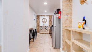 Un pasillo estrecho que conduce a una cocina con refrigerador, un comedor con mesa y sillas, y un ventilador fijado a la pared.