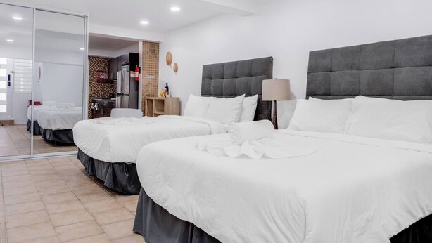 Habitación de hotel con dos camas, un espejo, un pequeño rincón americano y un baño visible a través de una puerta de vidrio.
