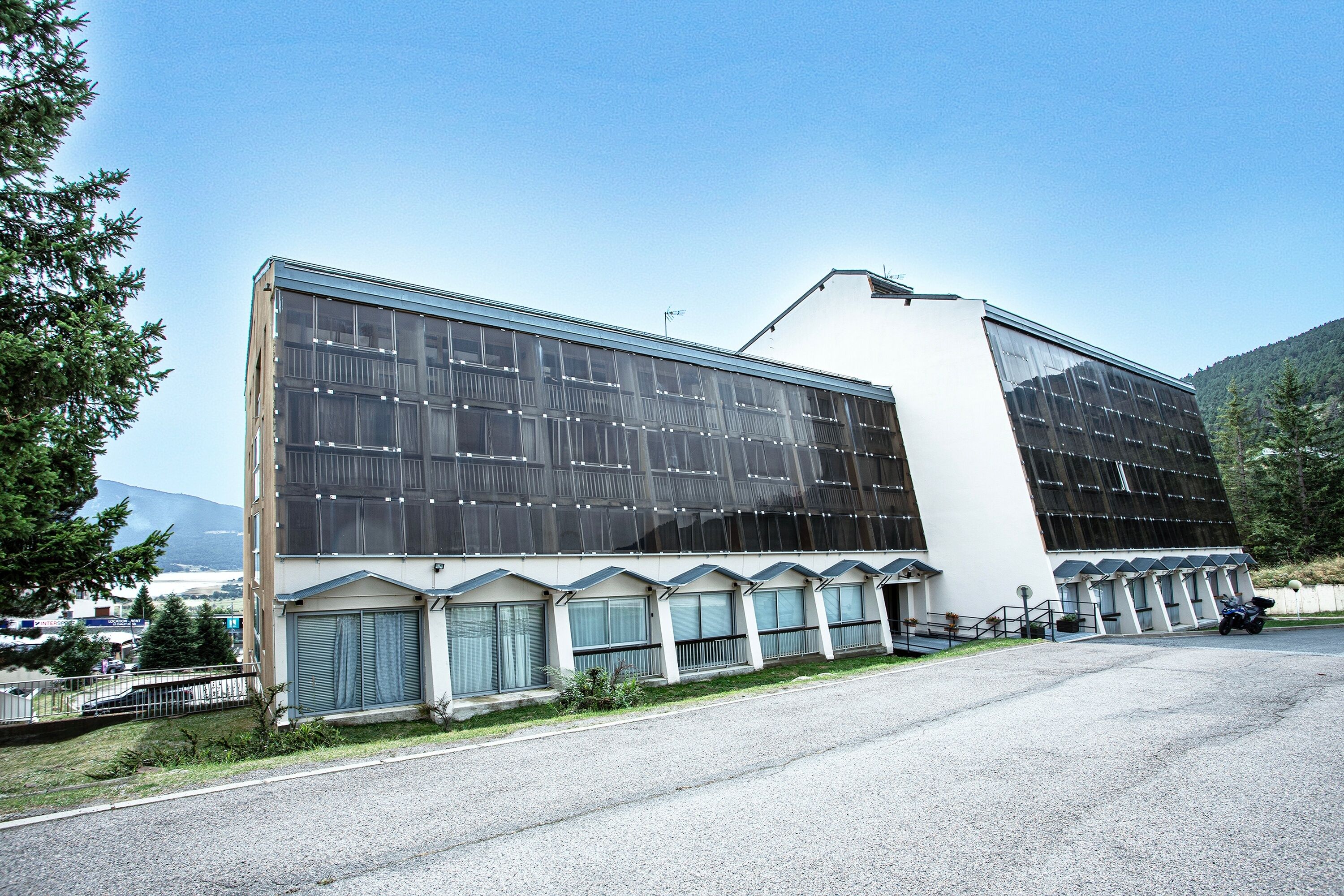 A modern building with large windows and a flat roof, surrounded by a parking area and greenery.