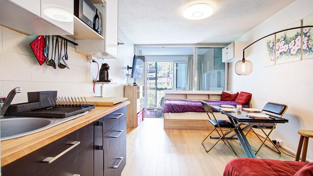 Cocina compacta con pileta, estufa y diversos utensilios. Se observa un comedor con mesa y sillas, junto a una cama al fondo.