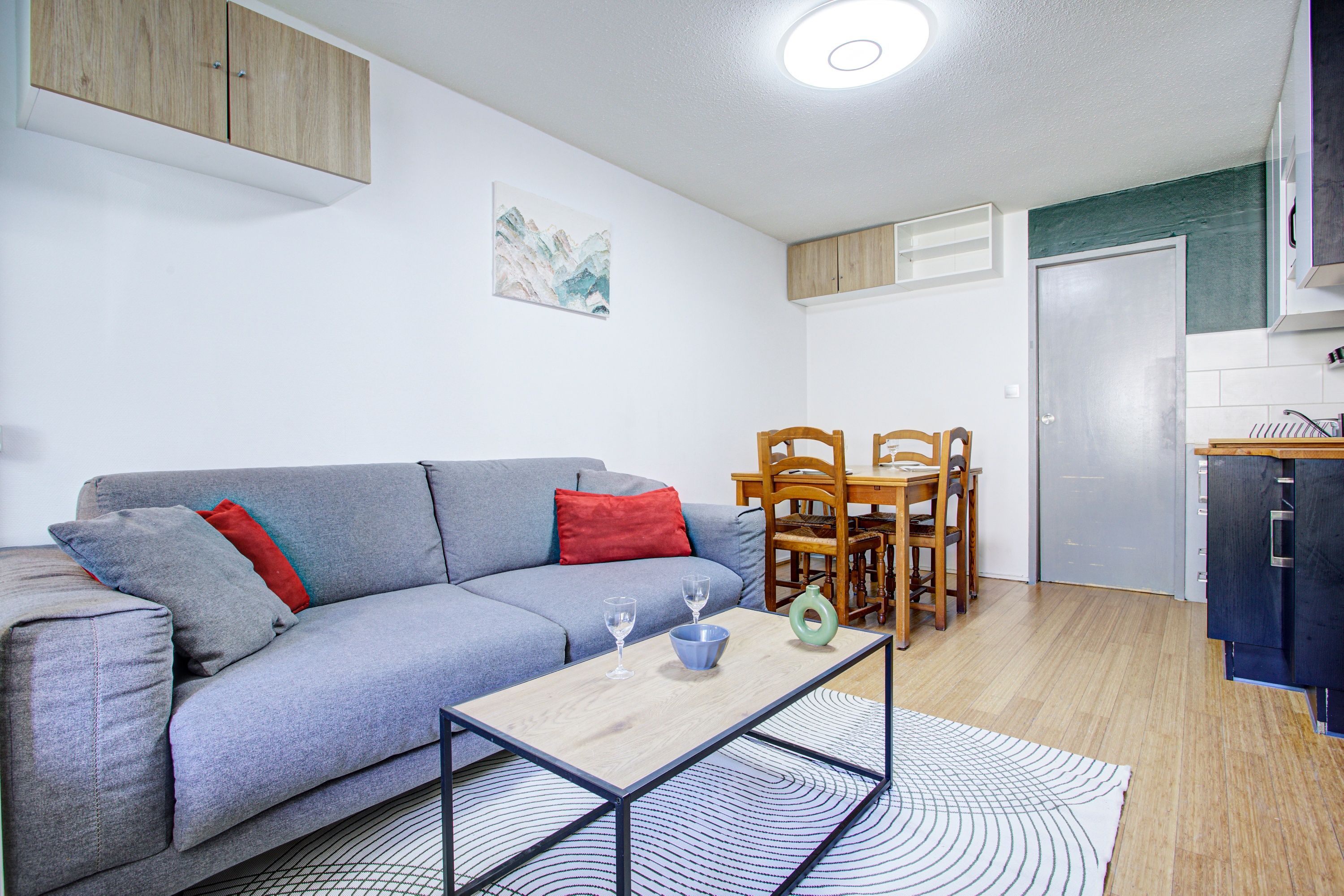 Un espai habitable compacte amb un sofà gris, taula de dinar amb cadires, zona de cuina i llum al sostre.