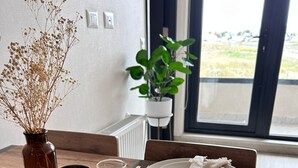 Une table à manger est dressée avec de l’argenterie dorée, des assiettes blanches et une nappe beige. On y voit deux verres contenant un liquide transparent, probablement de l’eau ou du vin, et un pot de couleur cuivrée. Un vase contenant des fleurs séchées est placé sur le côté gauche de la table.
