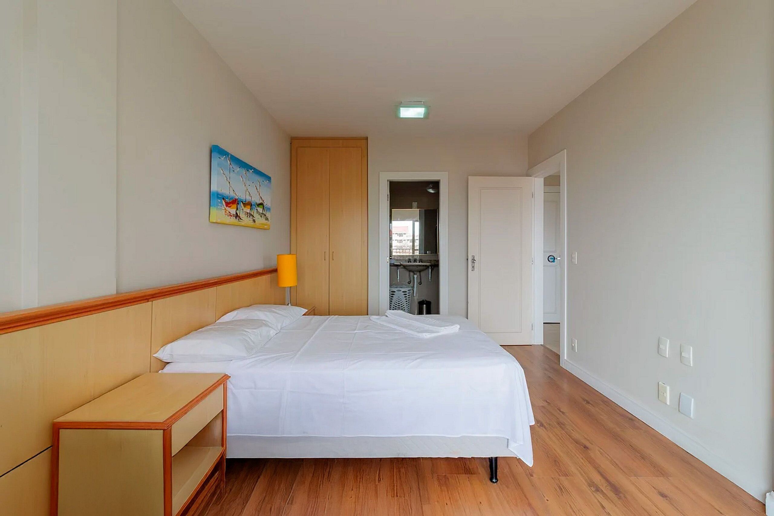 A hotel room with a bed, wooden headboard, a painting on the wall, a nightstand, and a door leading to a bathroom.