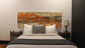 Una cama bien hecha con edredón gris y blanco, dos almohadas y una manta de color negro. Un cuadro abstracto moderno adorna la pared detrás de la cama. Dos mesitas de noche de madera flanquean la cama, cada una con una lámpara.
