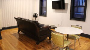 Una habitación con piso de madera, un sofá de cuero oscuro, una mesa redonda con sillas, un televisor instalado en la pared y dos ventanas.