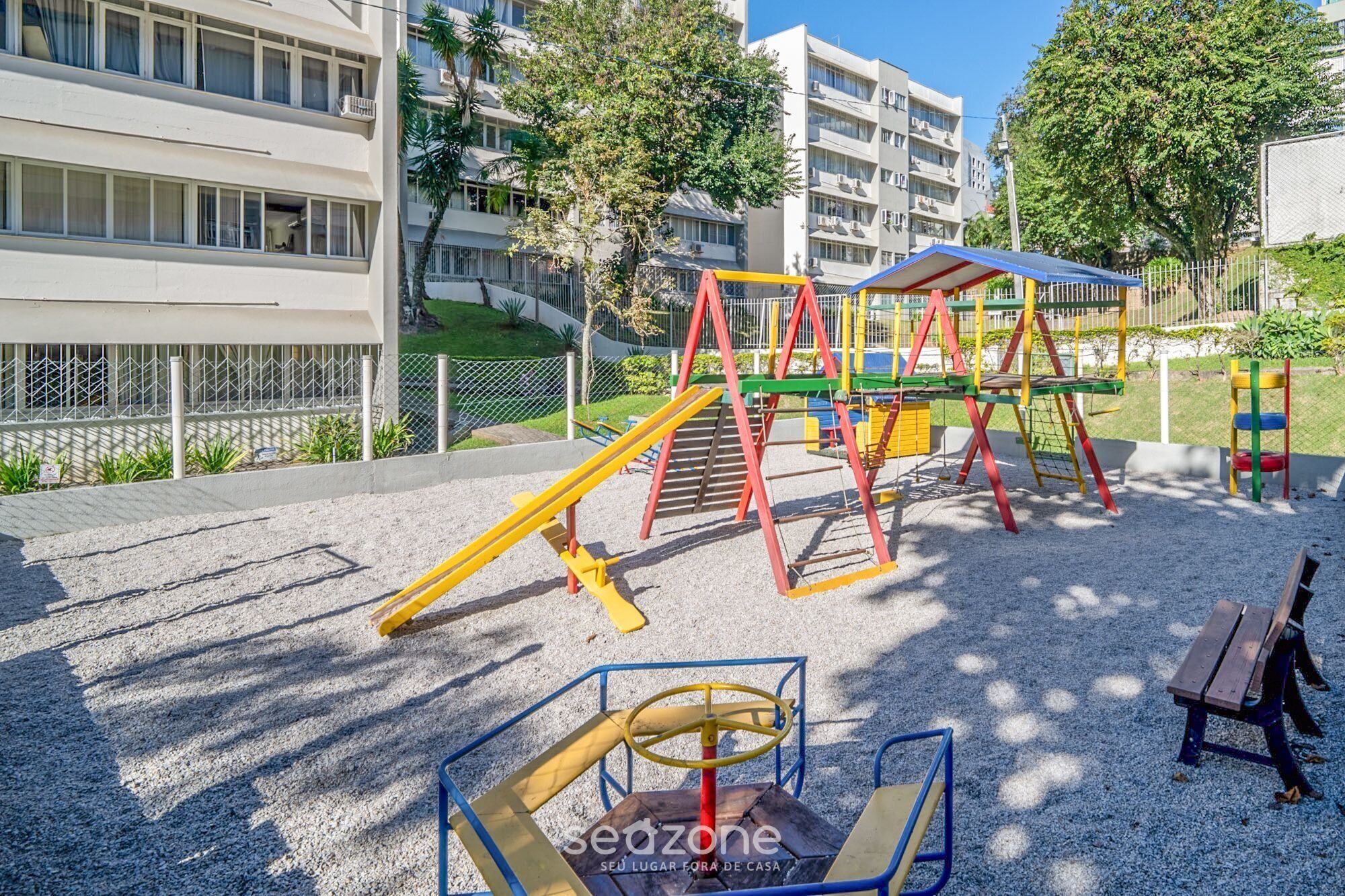 A playground with climbing structures and a seesaw, surrounded by a fence and buildings.