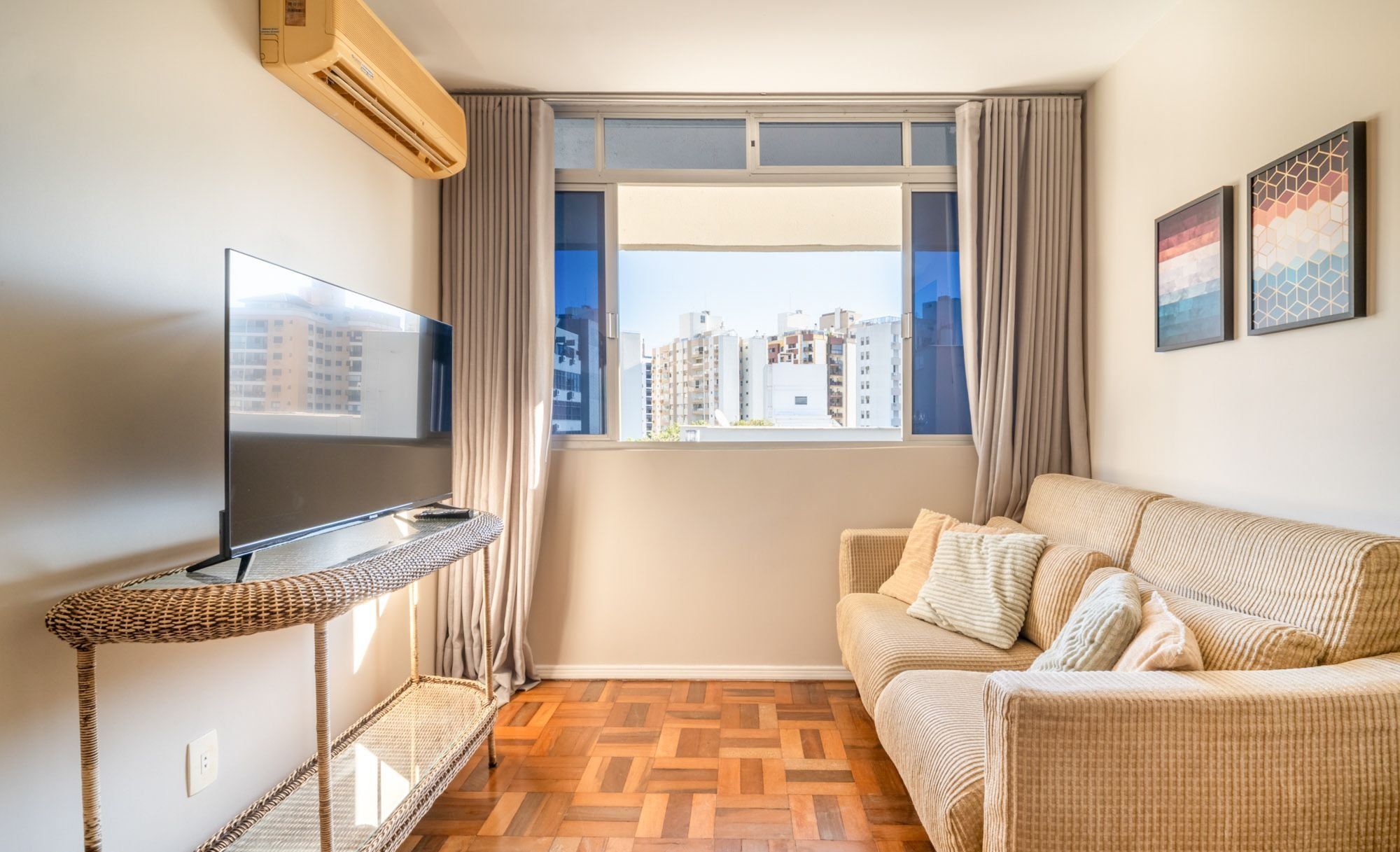 Ein Wohnzimmer mit Fernseher, einer Couch, einem Fenster mit Vorhängen und Blick auf Gebäude.