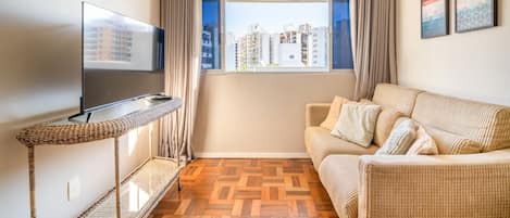 Ein Wohnzimmer mit Fernseher, einer Couch, einem Fenster mit Vorhängen und Blick auf Gebäude.