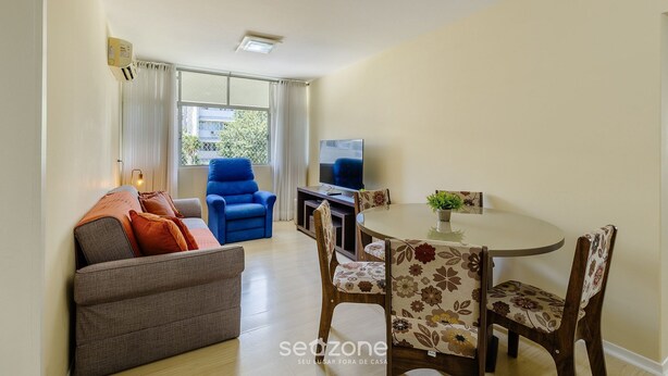 A living room with a sofa, a blue armchair, a dining table with floral chairs, a TV, and a window with curtains. - Apartment With Home Office in Floripa Jfm0062 (Florianópolis)