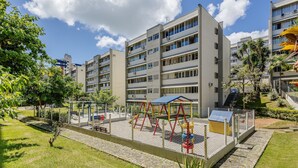 A playground area with swings and slides, surrounded by a fence, in front of a multi-story building with balconies. - Apartment With Home Office in Floripa Jfm0062 (Florianópolis)