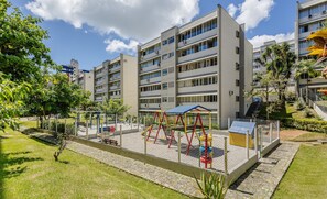 A playground area with swings and slides, surrounded by a fence, in front of a multi-story building with balconies. - Apartment With Home Office in Floripa Jfm0062 (Florianópolis)
