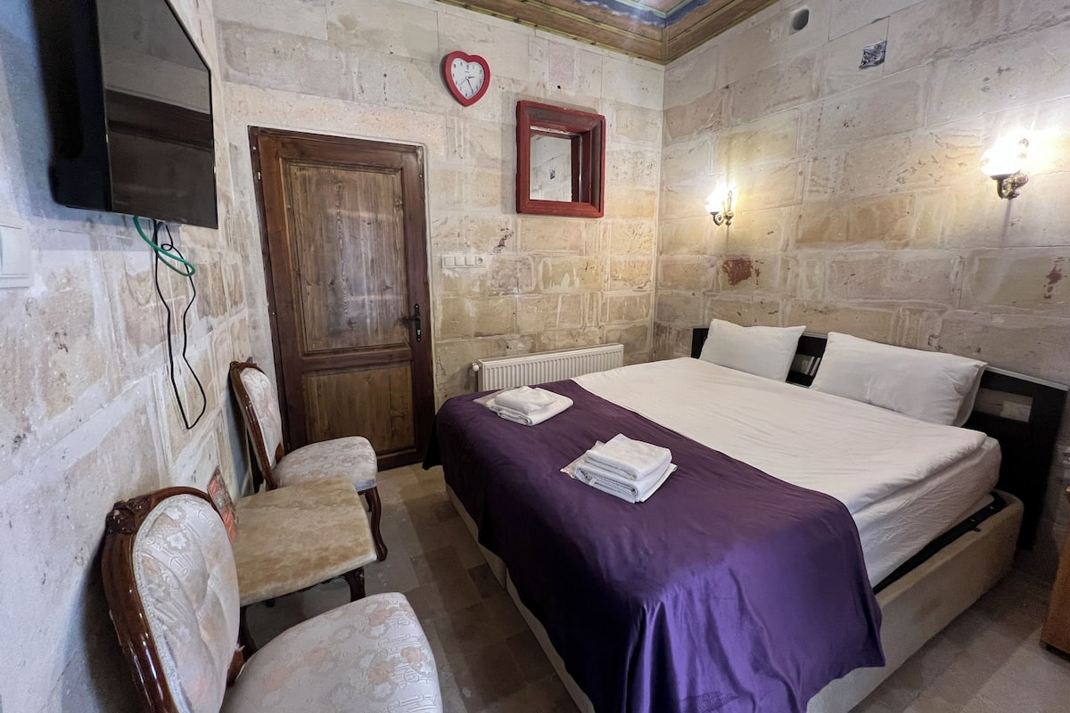 A room with a bed, a wooden door, a chair, a television, and stone walls.
