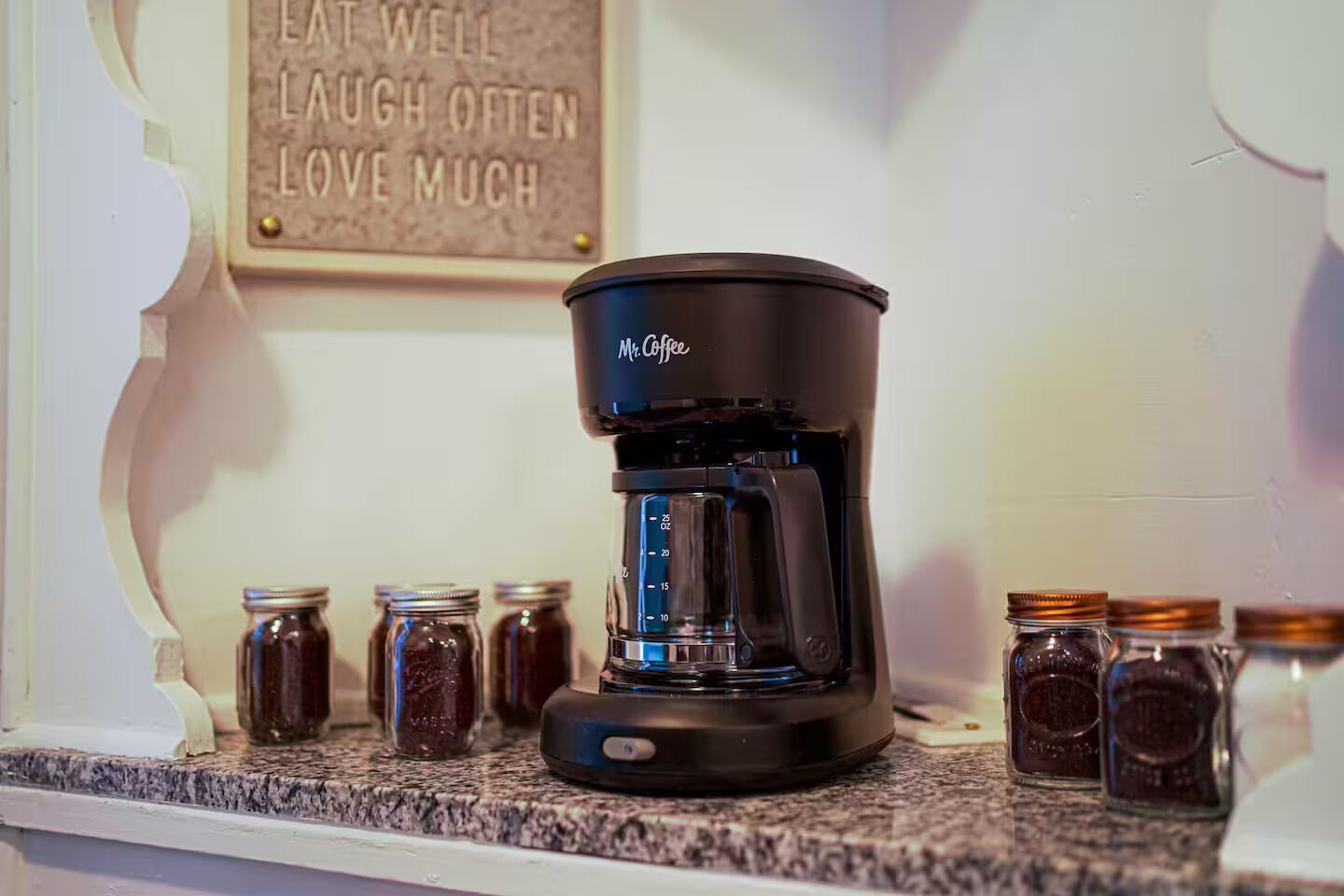 Una cafetera sobre una encimera de cocina con frascos de especias y una frase motivacional en la pared.