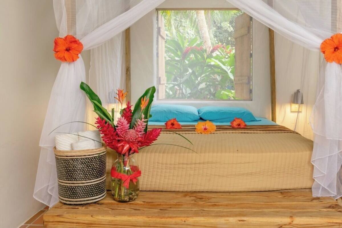 A room with a bed, a window with curtains, a vase with flowers, and a woven basket.