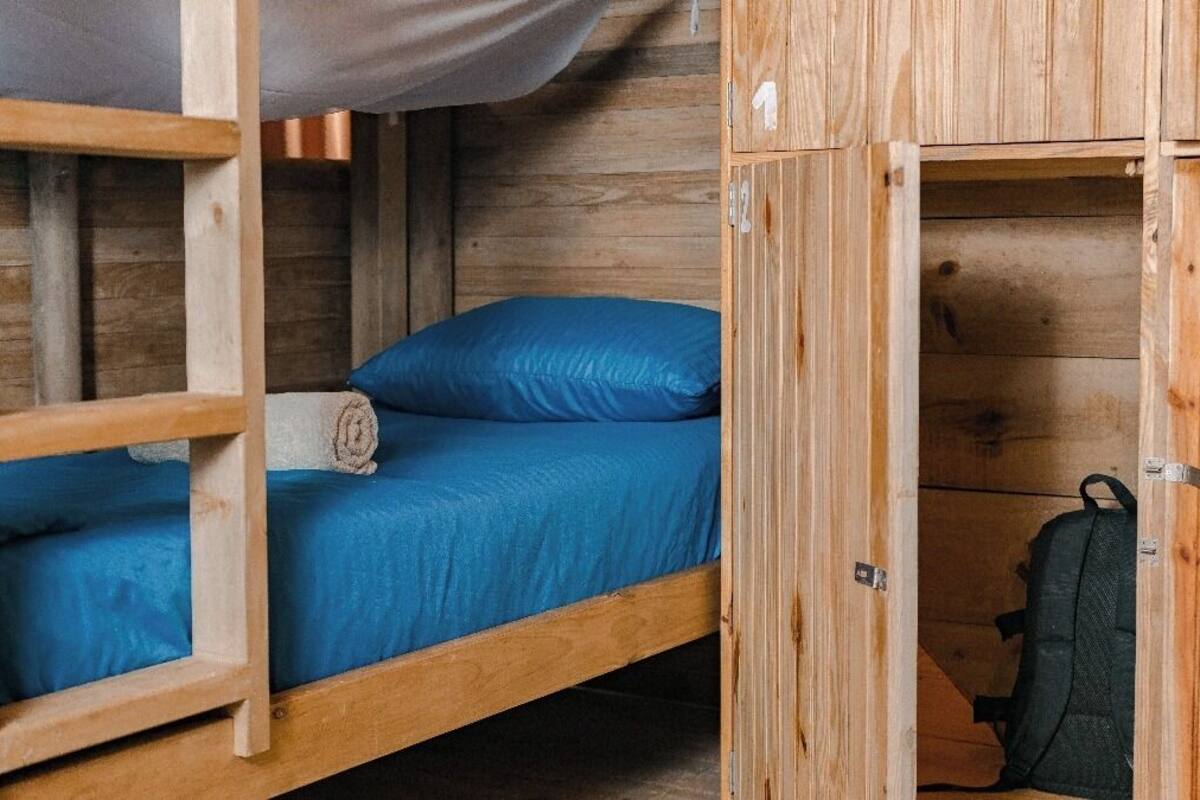 A wooden bunk bed with a blue mattress and pillows, a wooden wardrobe with a suitcase inside, and a wooden ladder.