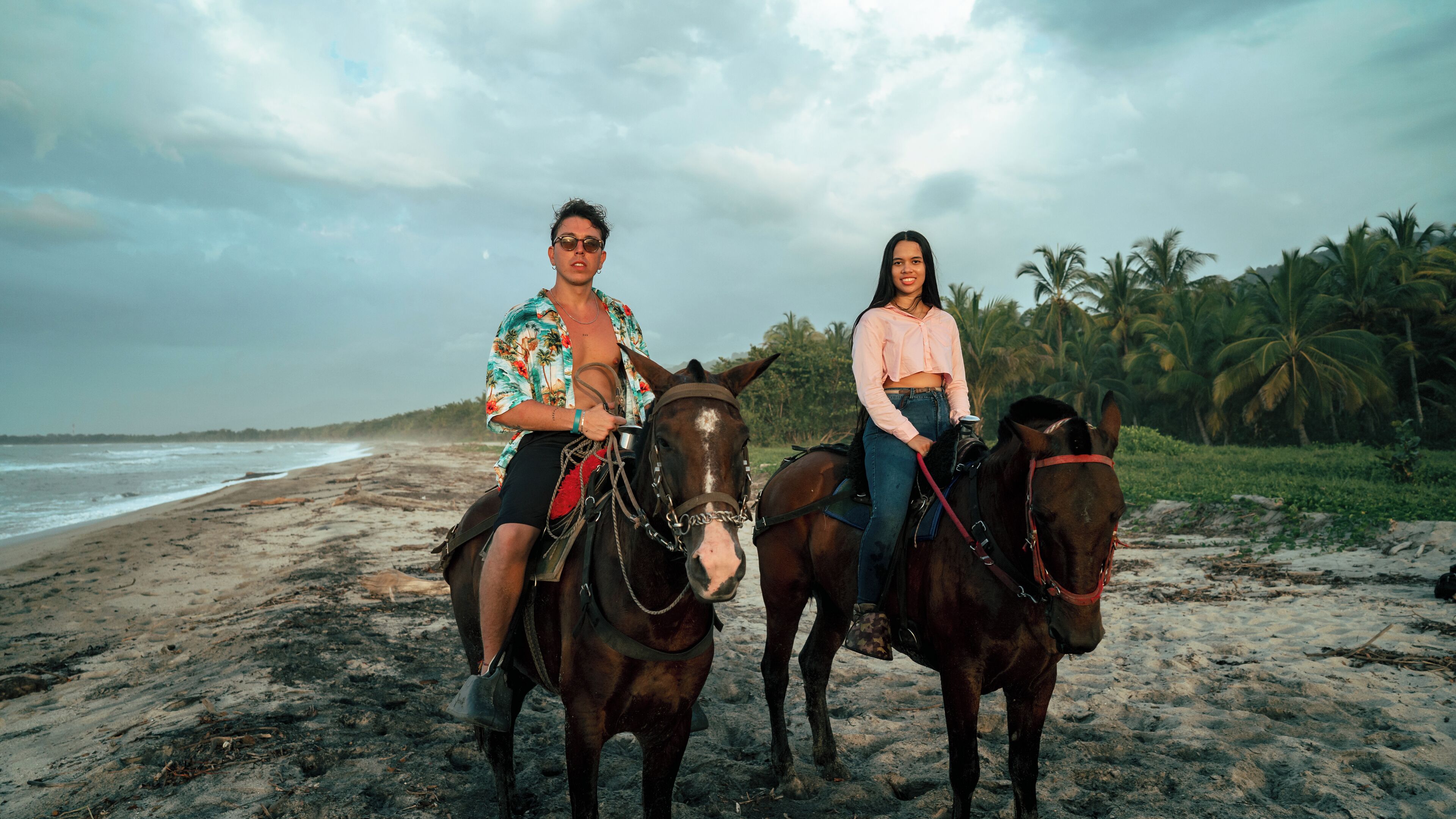 Two people on horseback on a beach with palm trees and the ocean in the background.