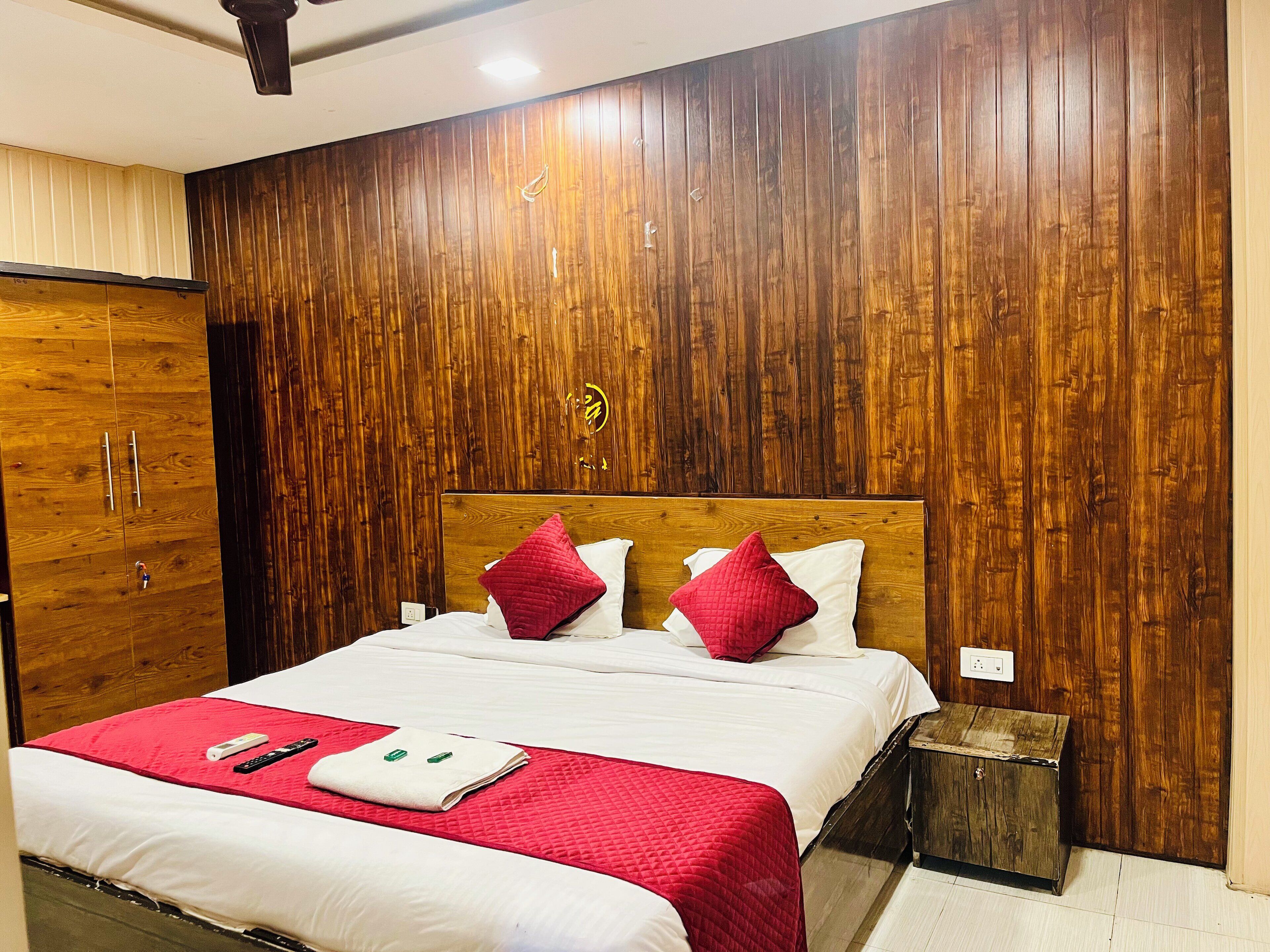 A hotel room with a wooden panel wall, a bed with white bedding and red pillows, a wooden nightstand, and a red quilted blanket.