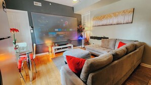 A modern living room with a grey sofa, a coffee table, and a TV mounted on the wall displaying streaming services. - Dream / King Bed / 120" Projector / Canopy Bed (Philadelphia)