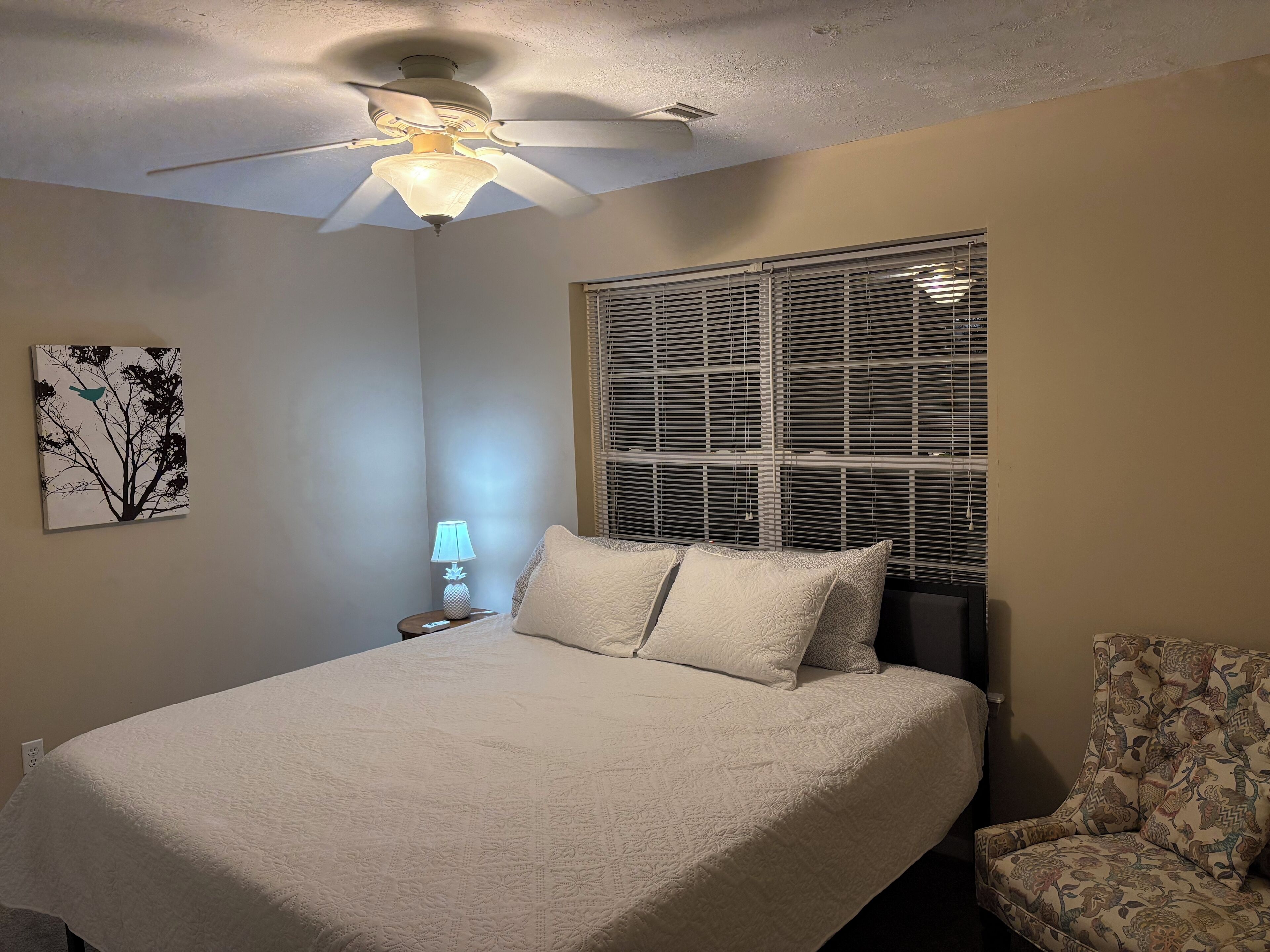 A bedroom with a bed, a ceiling fan, a window with blinds, a nightstand with a lamp, and a chair.