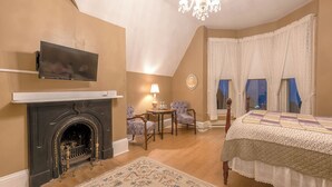 Un dormitorio con chimenea, un televisor montado encima, una cama con colcha, dos sillas, una mesita y una ventana con cortinas.