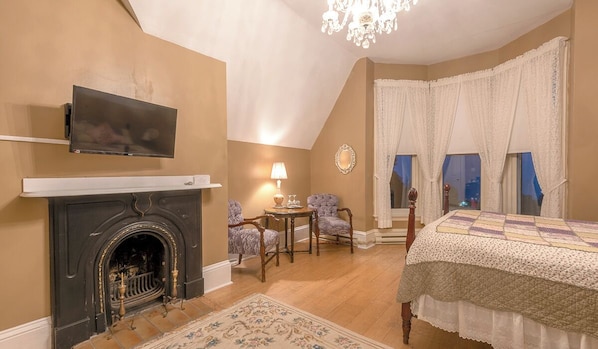 Un dormitorio con chimenea, un televisor montado encima, una cama con colcha, dos sillas, una mesita y una ventana con cortinas.