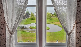Janela com cortinas brancas, vista de área gramada com uma pequena ponte e lago.