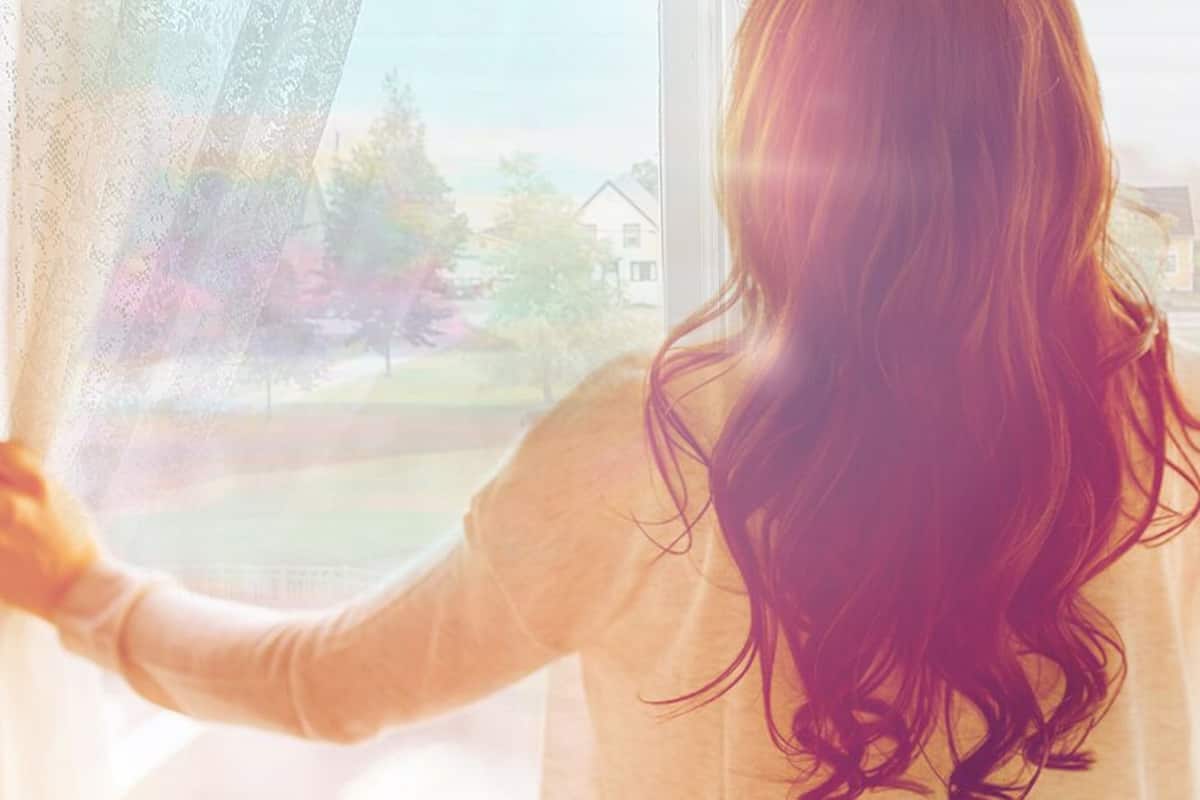 Una persona de cabello largo mirando por una ventana con cortinas translúcidas.