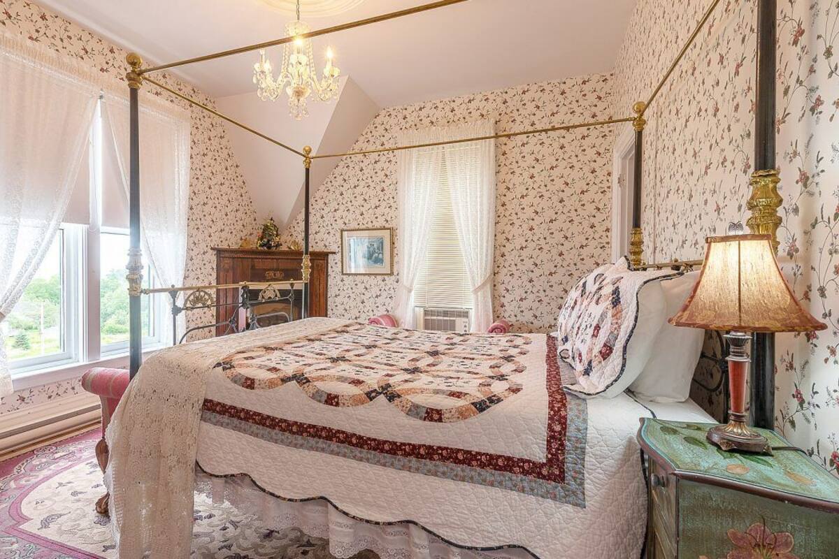 Un dormitorio con cama con dosel, papel tapiz floral, una araña y un aparador de madera, además de una mesita de noche con lámpara.