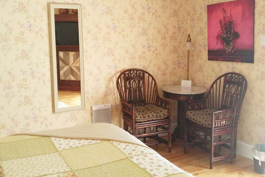 A room with floral wallpaper, a bed with a quilt, two wooden chairs, a small round table, and a painting on the wall.