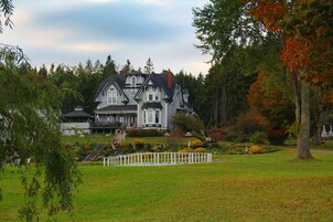 Uma casa grande e histórica, com uma varanda frontal imponente, rodeada por um jardim bem cuidado e uma cerca branca.