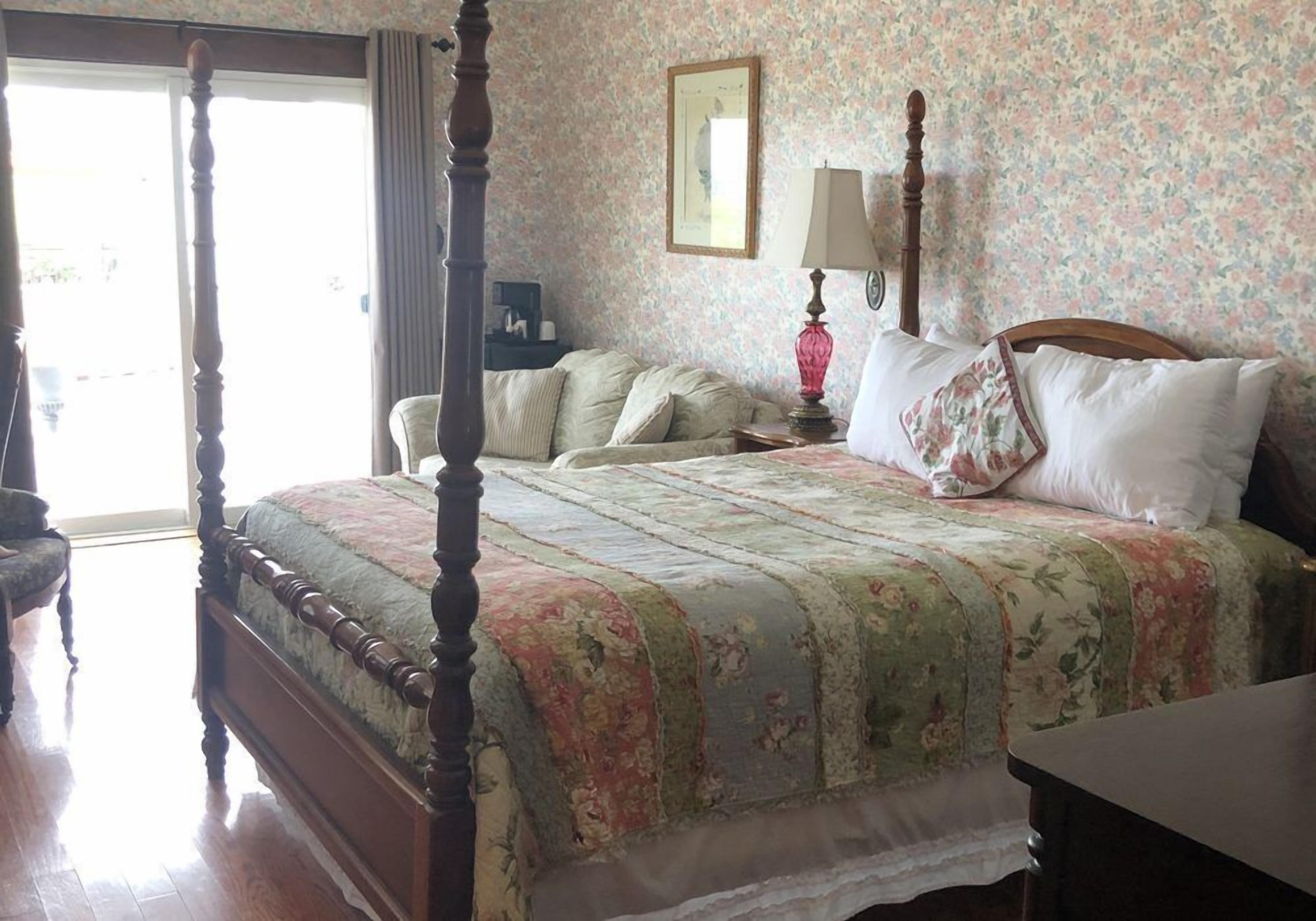 Una cama con dosel con ropa de cama floral, una mesita de noche con lámpara y una silla en una habitación con papel tapiz de flores.