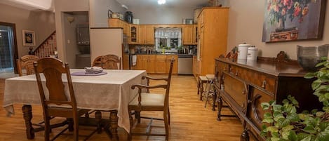 A dining area with a wooden table and chairs, a chandelier, and a kitchen in the background.