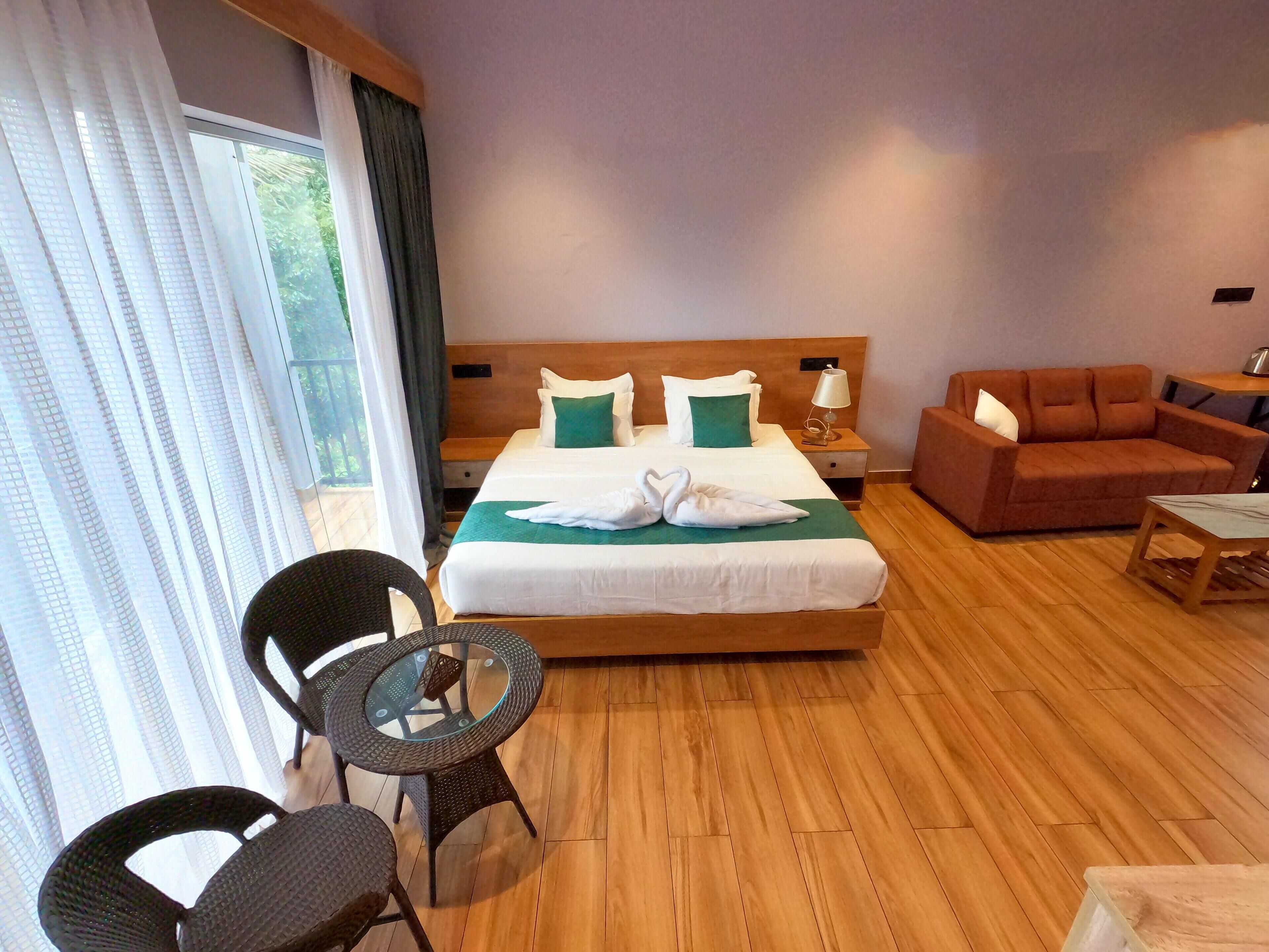 A hotel room with a large bed, a wooden headboard, a green throw, two white towels shaped like swans, a brown sofa, a round glass table, and a wicker chair.