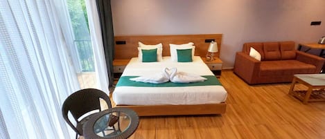 Ein Hotelzimmer mit einem großen Bett, einem hölzernen Kopfteil, einer grünen Decke, zwei weißen Handtüchern in Schwanenform, einem braunen Sofa, einem runden Glastisch und einem Korbstuhl.