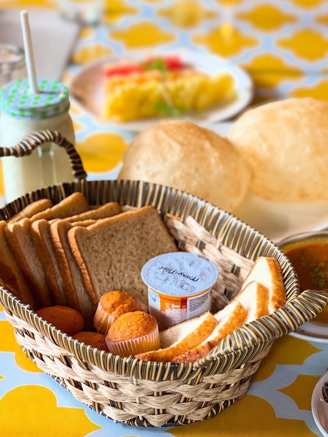 A wicker basket containing slices of bread, muffins, and a container of yogurt.