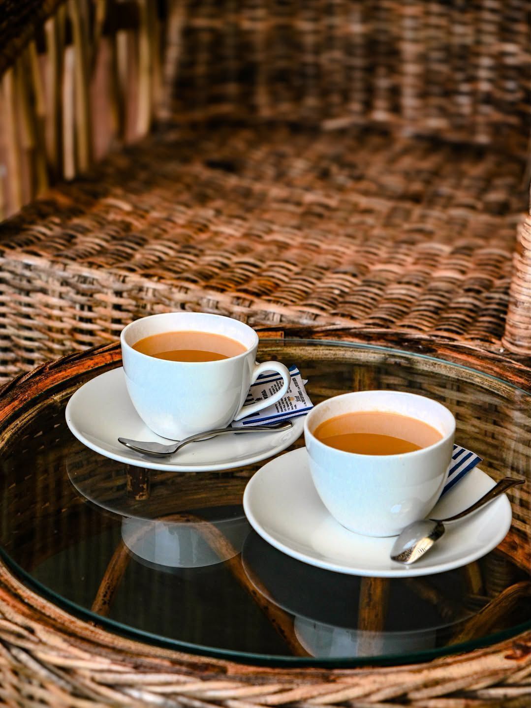 Dos tazas blancas de té sobre una bandeja de cristal con platitos y cucharas, sobre una silla de mimbre.