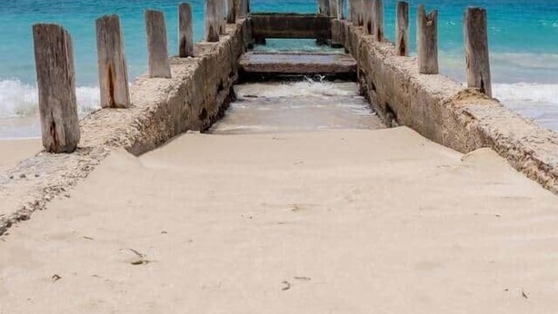 Une jetée en béton qui mène à une plage bordée d’une eau turquoise et cristalline.