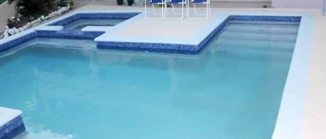 A rectangular swimming pool with a tiled edge, a small platform with four blue chairs, and potted plants around the pool area.