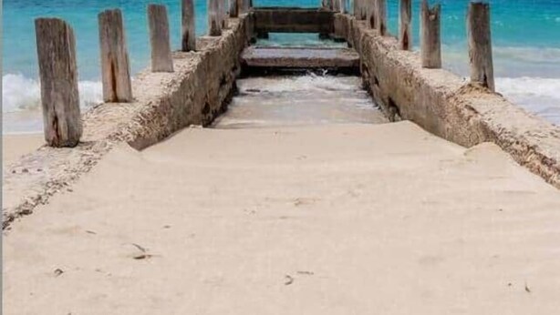 Een betonnen steiger die naar een strand leidt, met helderblauw water en een heldere lucht.