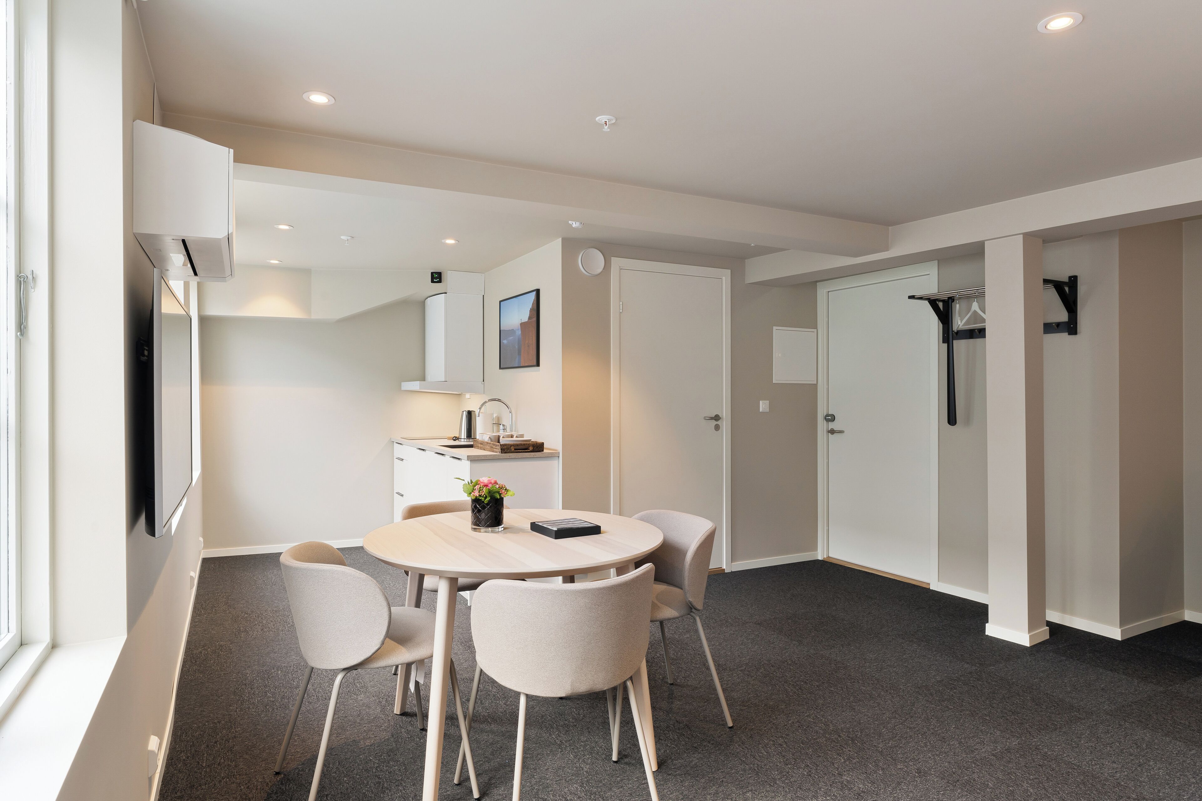 A modern hotel room with a round dining table, four chairs, a small kitchenette, a large window, and a door leading to a bathroom.
