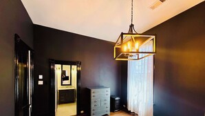 A bedroom with a wooden floor, a bed, a dresser, a mirror, and a hanging light fixture.