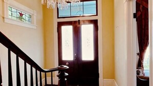 A wooden staircase leading to a dark wooden door with a window. A chandelier hangs from the ceiling, and a bench is placed at the bottom of the stairs.