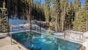 Une piscine aux eaux limpides, entourée d’une clôture métallique, avec un sol recouvert de neige et de grands arbres en arrière-plan.