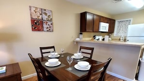 A dining area with a wooden table set for four, surrounded by chairs. There is a small side table with a box on it. The room has wooden flooring and a ceiling light fixture.