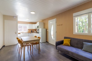 A modern kitchen with white cabinets, a dining table with chairs, a sofa with cushions, and a window with a view of trees. - Authentic Stay in Dordogne – Comfortable Gîte with Heated Pool (Thenon)