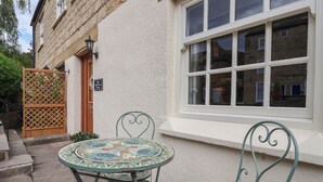 Un patio con una mesa redonda y dos sillas, una puerta de madera, y un edificio con fachada blanca y una ventana.