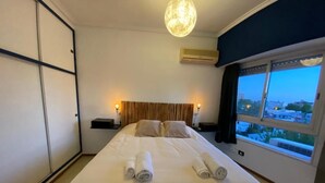 A hotel room with a large bed, two pillows, a headboard, a nightstand, a lamp, and a window with a view of buildings.