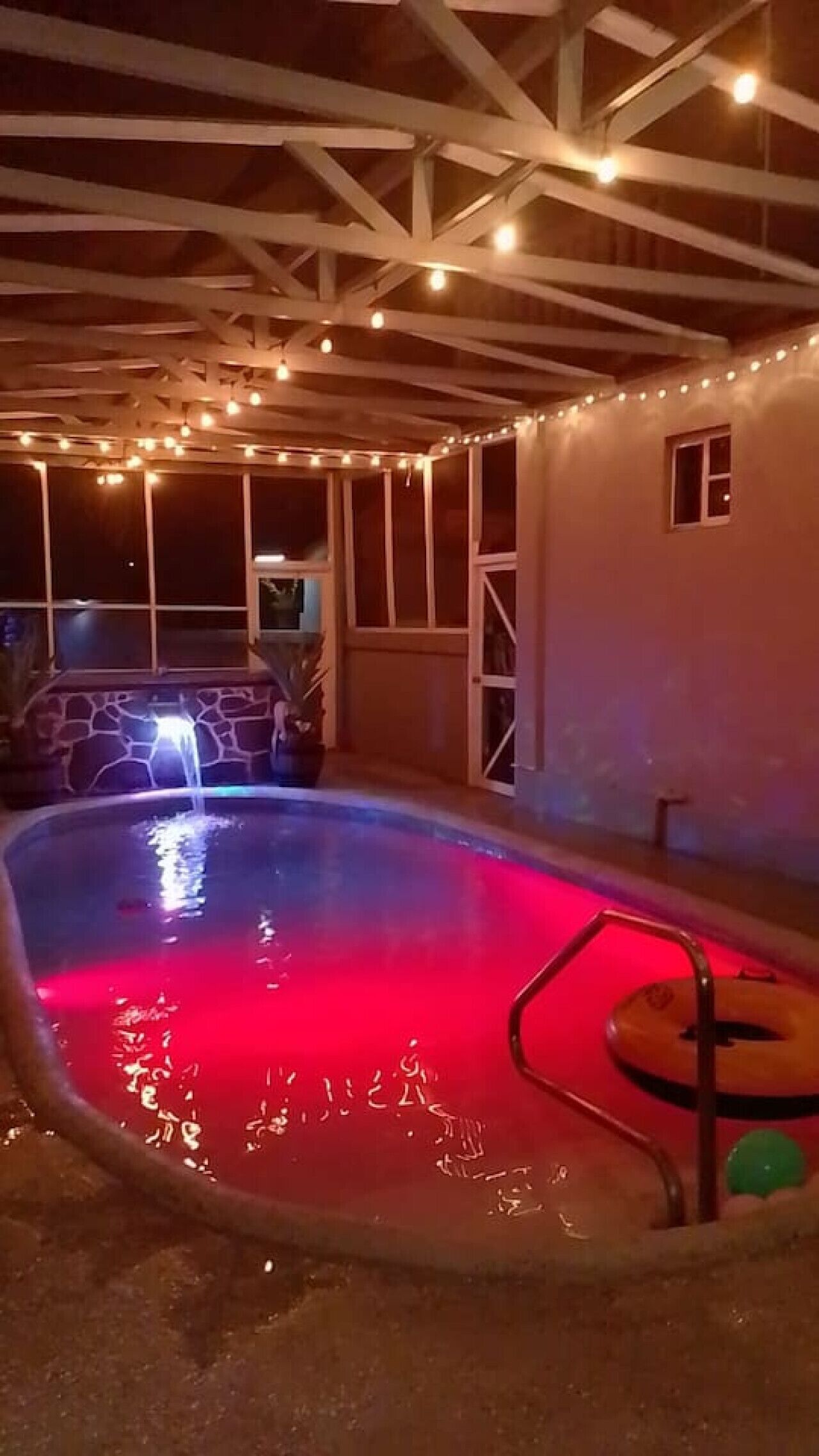 Una piscina con una luz roja, rodeada por un espacio techado con vigas de madera y luces colgantes.
